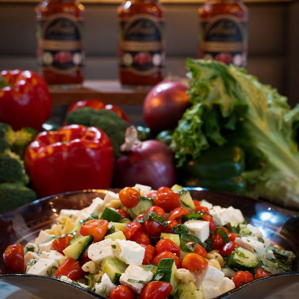 Pasta salad with tomatoes, cucumbers, and feta cheese, surrounded by fresh vegetables and sauce jars.