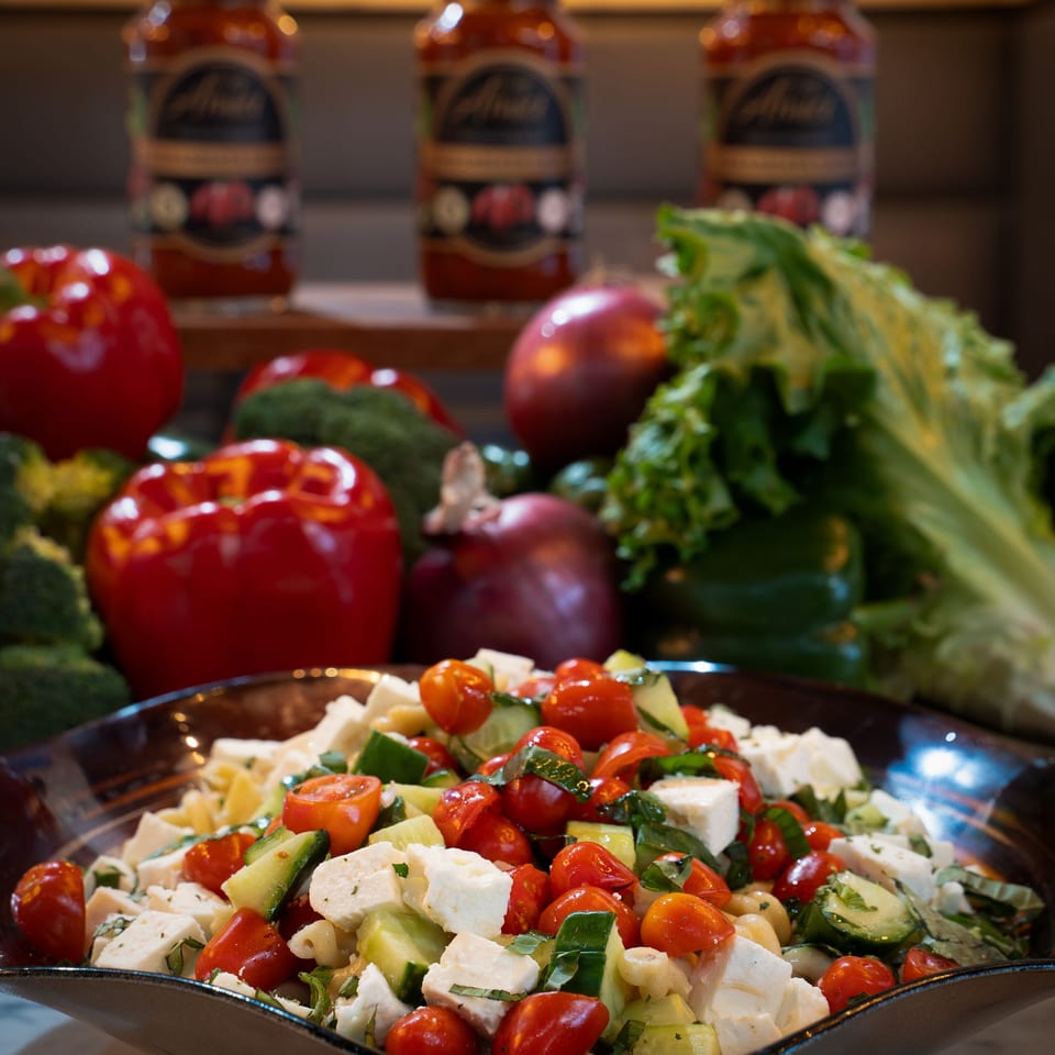 Pasta salad with tomatoes, cucumbers, and feta cheese, surrounded by fresh vegetables and sauce jars.